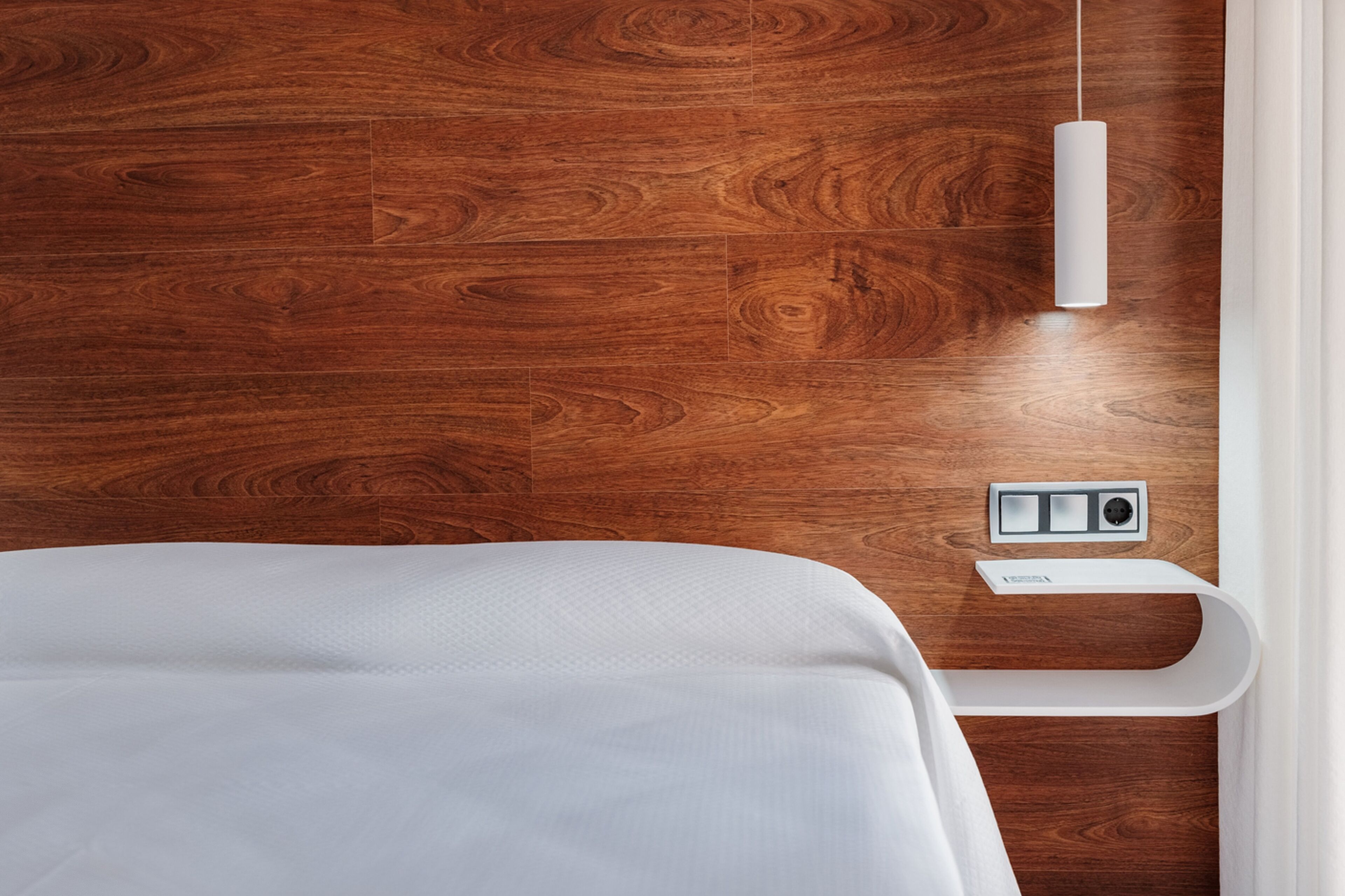 Een slaapkamer met een houten vloer, een wit bed, een nachtkastje met stopcontact en een hanglamp.