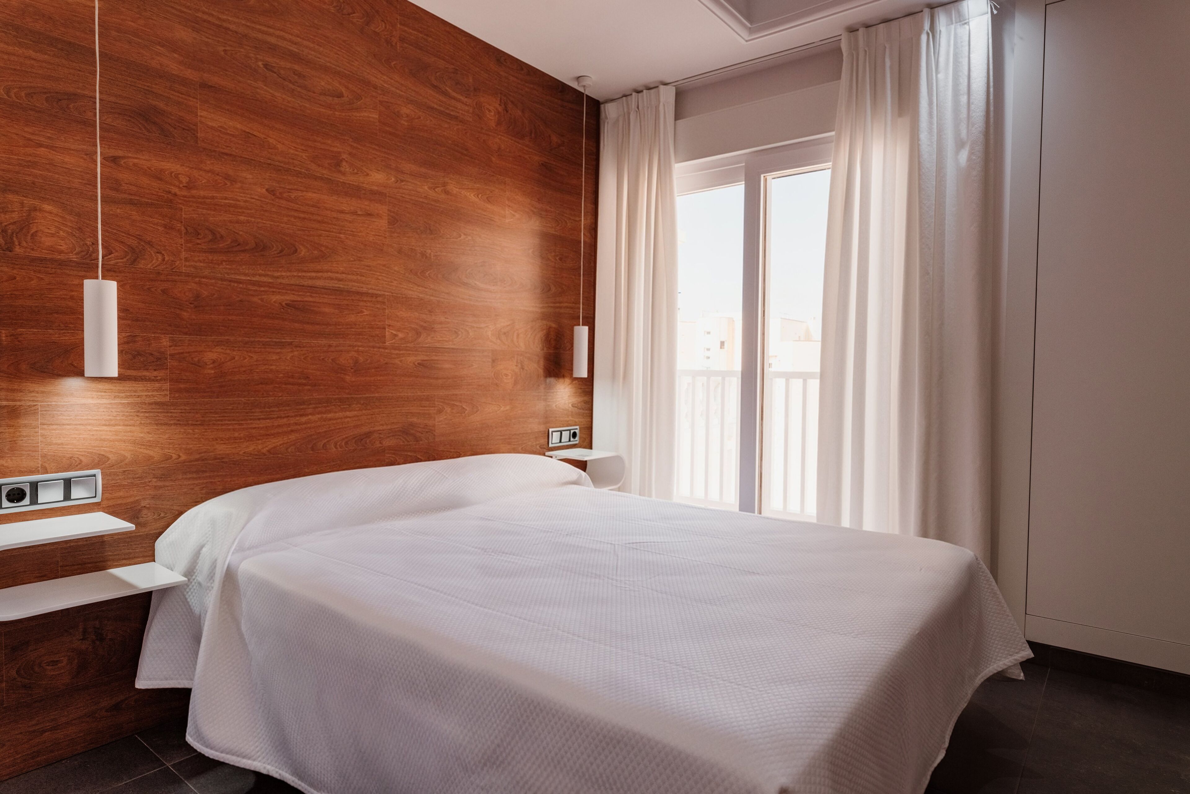 A modern hotel room with a wooden accent wall, a large bed, a bedside table, and a view of the city through a window with curtains.