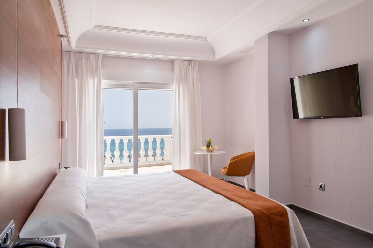 A hotel room with a bed, a TV, a small table with a plant, and a view of the sea through large windows.