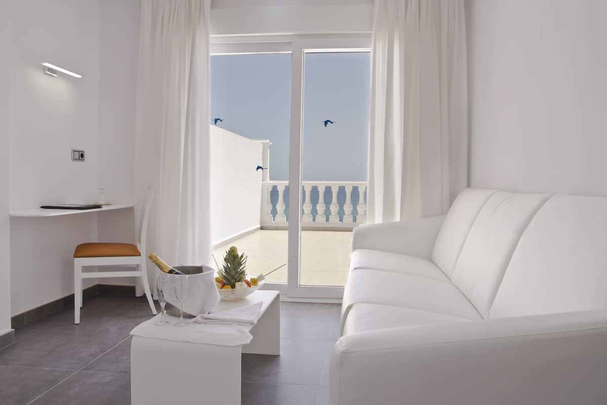 A modern, white-themed living room with a white sofa, a white table with a bottle and glasses, a white chair, and a balcony with a railing.