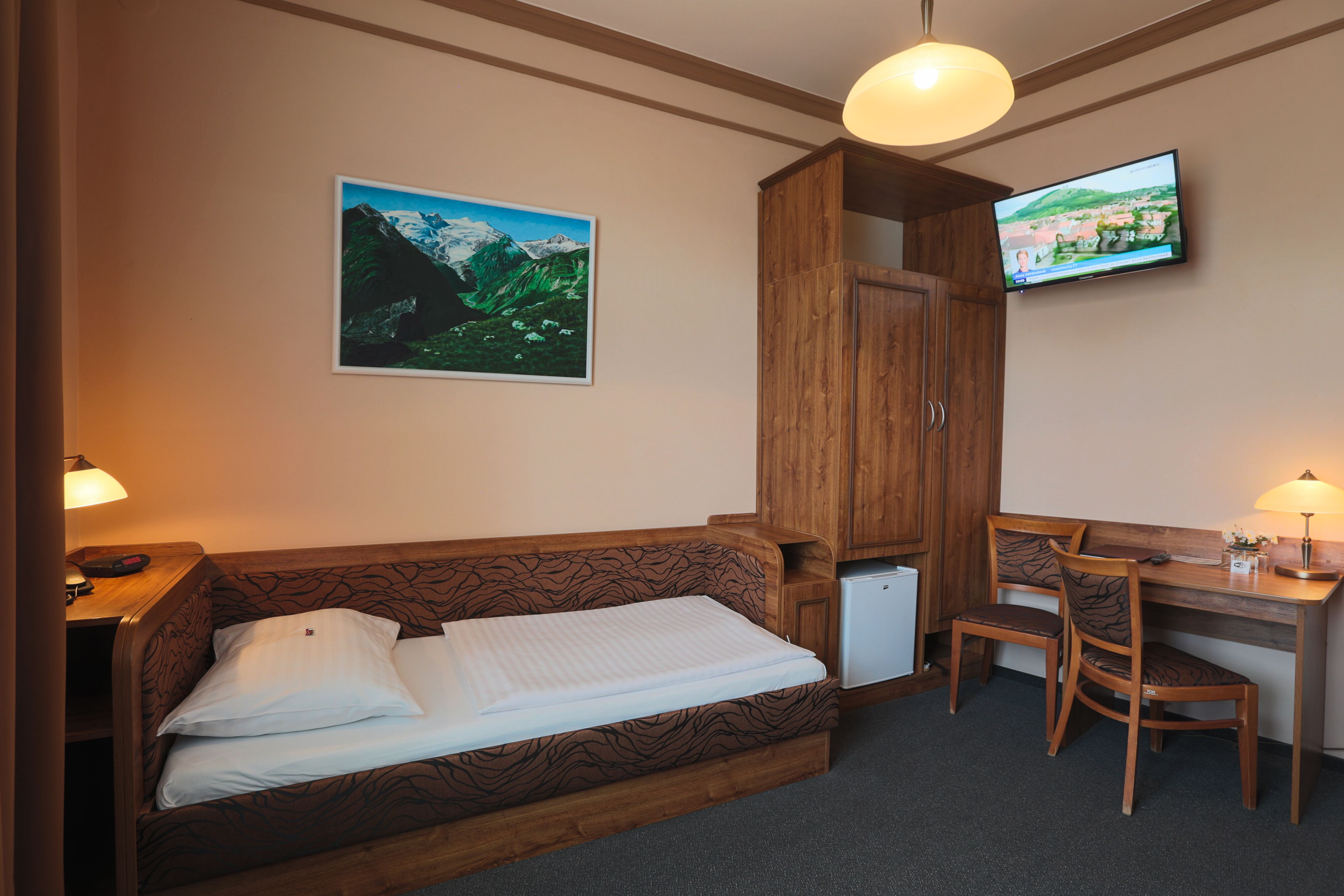 A room with a wooden sofa bed, a desk with a chair, a television, and a picture of a mountain landscape on the wall.