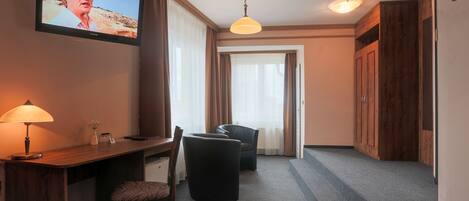 Habitación de hotel con televisor montado en la pared, escritorio con lámpara, una silla y un sofá. Se observa un armario de madera y una puerta que da a otra habitación.