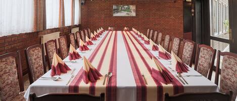 A long dining table set with red napkins, white tablecloth, and floral-patterned chairs in a brick-walled room with a window and a door.
