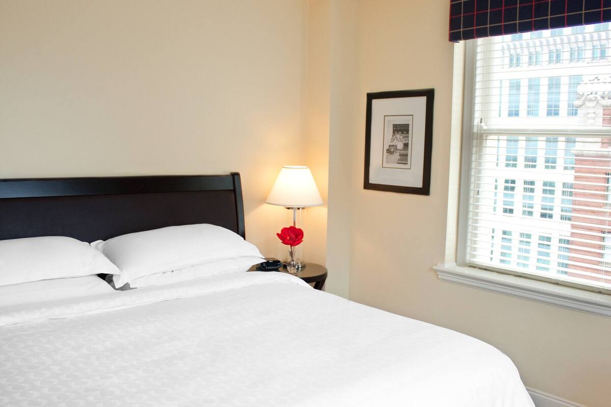 A neatly made bed with white linens, a bedside lamp, and a framed picture on the wall.