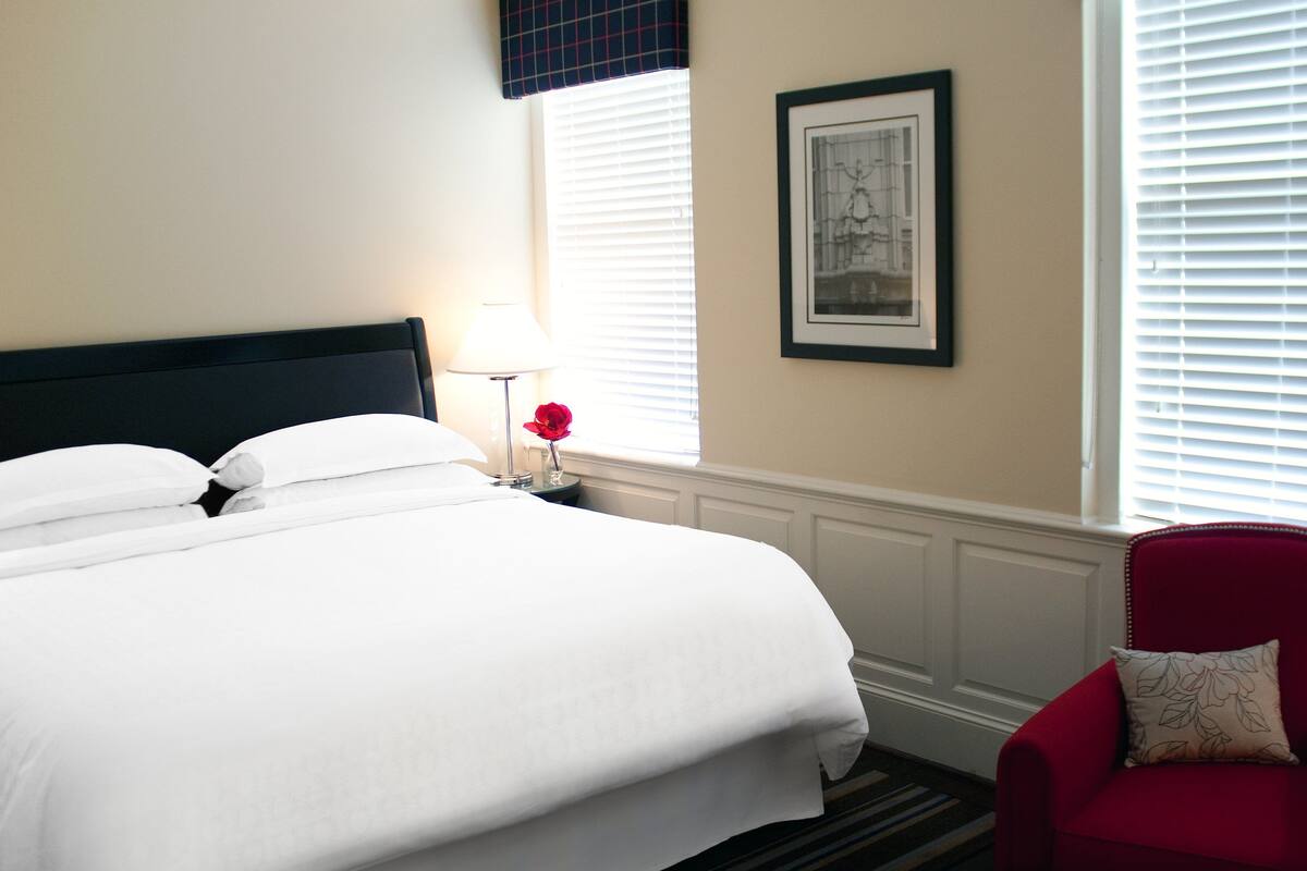 A neatly made bed with white linens, a red chair with a patterned pillow, a bedside table with a lamp, and a framed picture on the wall.