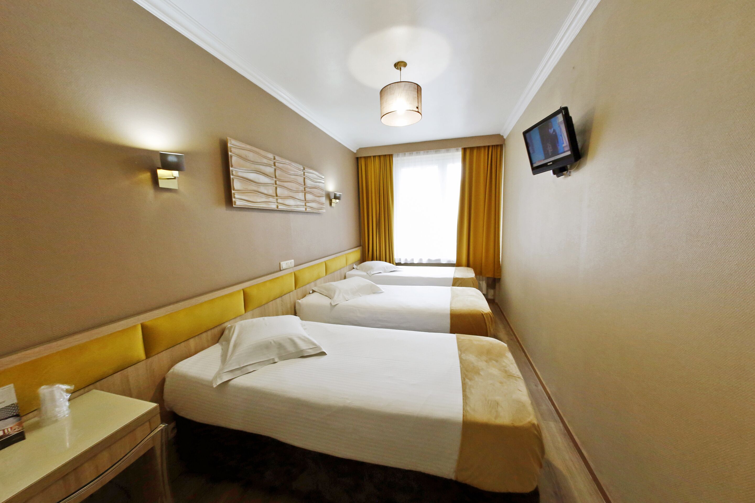 A hotel room with three beds, a television, a window with curtains, and a wall-mounted light fixture.