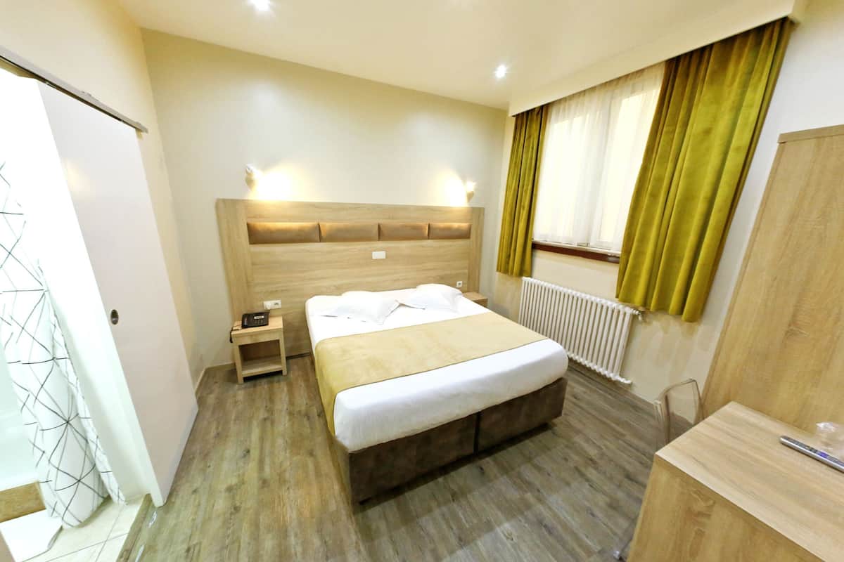 Ein Schlafzimmer mit Holzkopfende, Nachttisch, Bett mit weißer Bettwäsche, ein weiterer Nachttisch und ein Fenster mit Vorhängen.