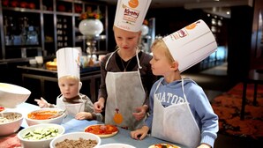A group of people wearing chef hats and aprons, preparing food at a table with various ingredients and bowls.