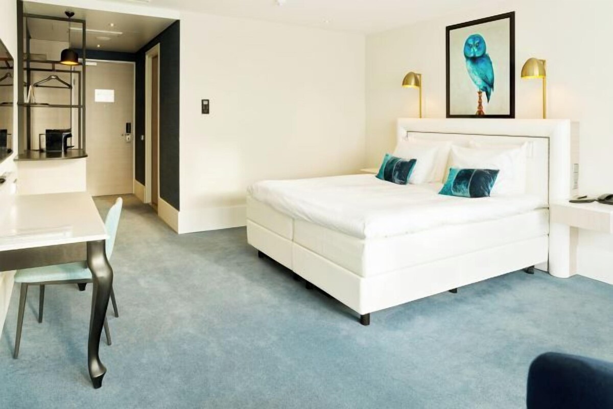 A modern hotel room with a white bed, a desk, and a chair. There is a painting of an owl on the wall.