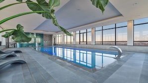 An indoor swimming pool with clear blue water, surrounded by large windows offering a city view.