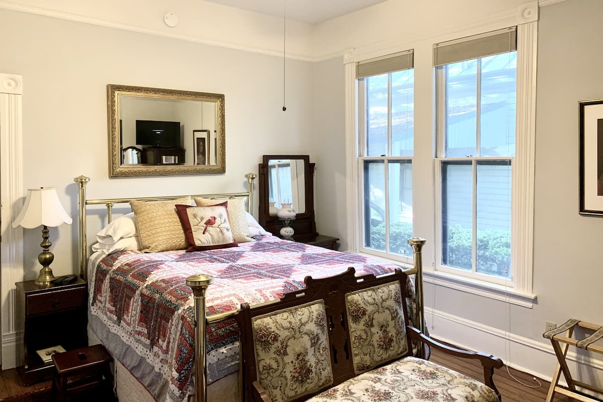 Une chambre à coucher avec un lit, une table de chevet, une chaise, un miroir et deux fenêtres.