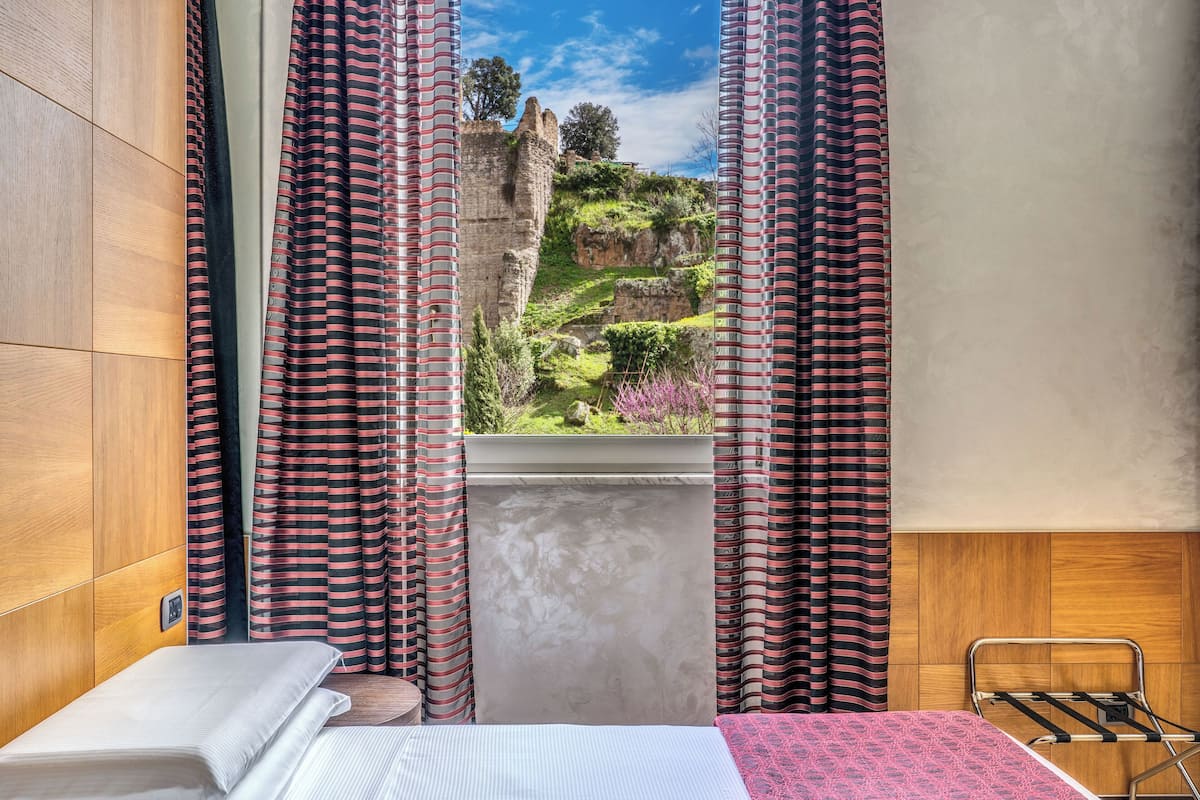 Una habitación con una cama, un ventanal con cortinas y vistas a un castillo y follaje.