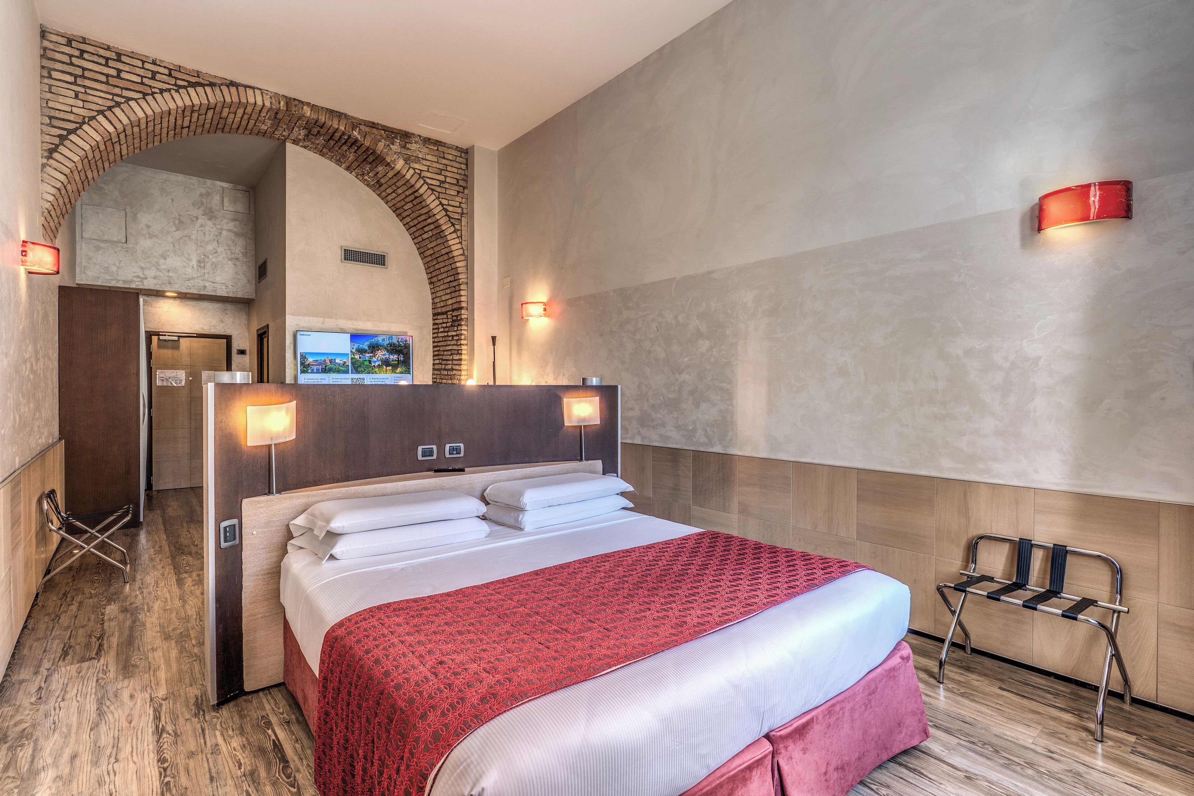 A hotel room with a large bed, a red patterned bedspread, a wooden bench, and a brick archway.