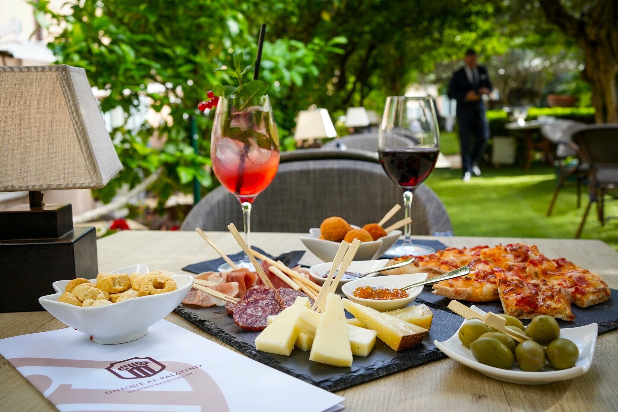 Et bord med et udvalg af oste, charcuteri, oliven og pizza, serveret med et par glas rødvin og en cocktail, udendørs med grøn vegetation i baggrunden.