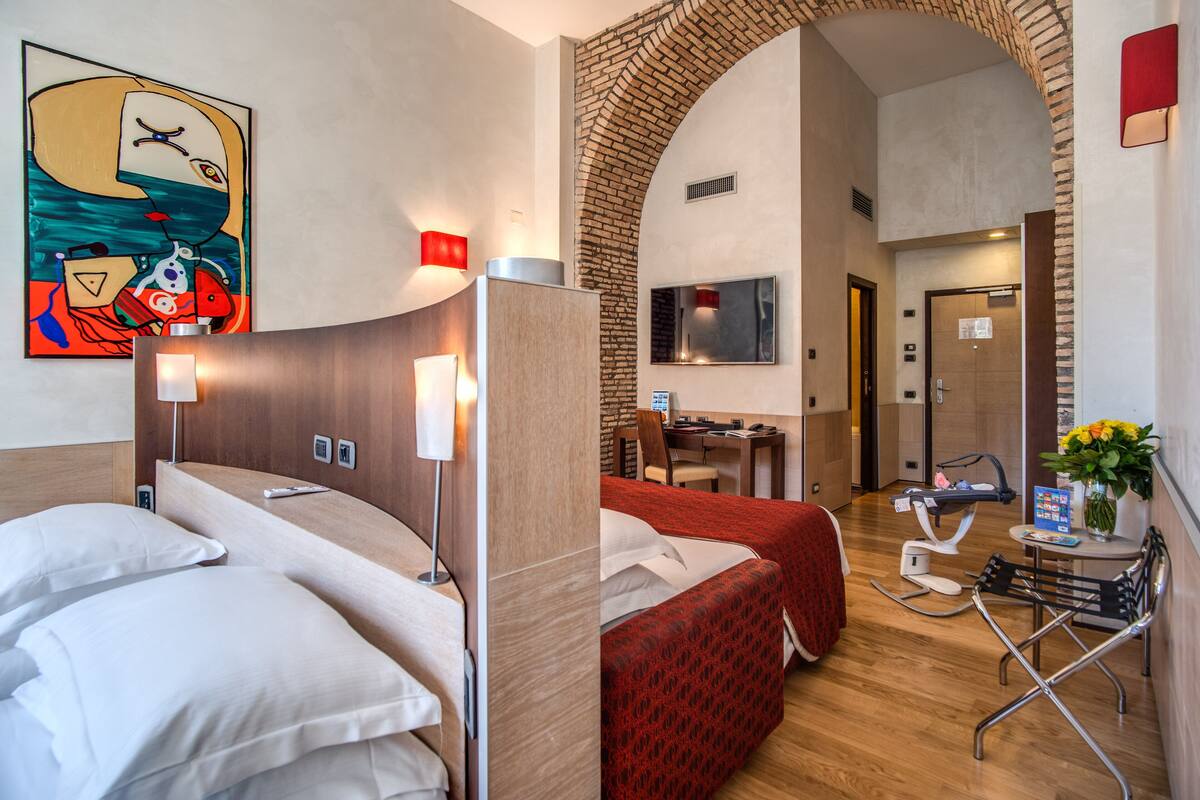 Habitación de hotel con dos camas, un escritorio, una silla, un televisor y un baño visible a través de un arco.