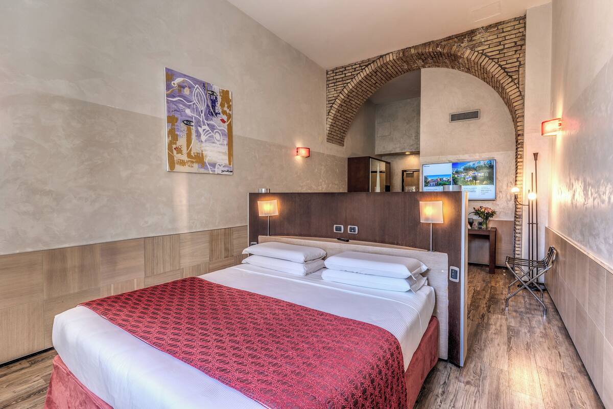 Habitación de hotel con una cama grande, cabecera de madera, colcha estampada de color rojo, televisor montado en la pared y un arco de ladrillo.