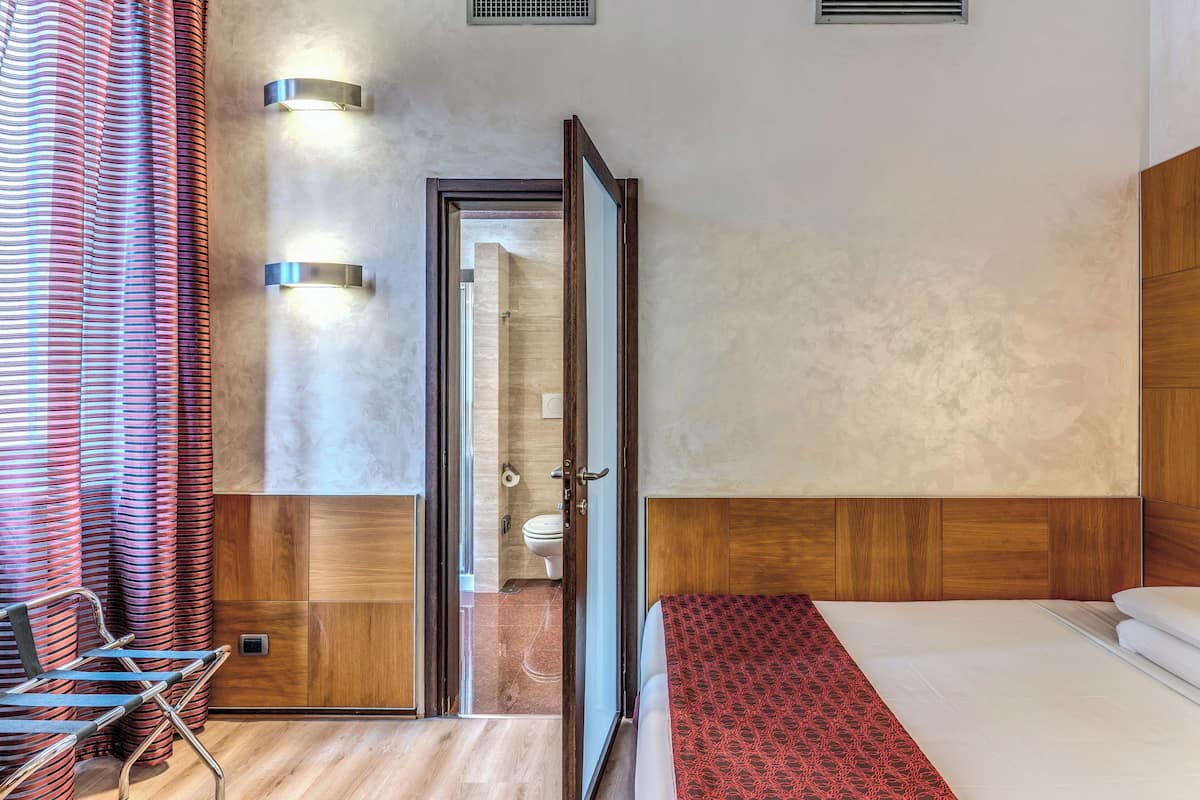 Habitación de hotel con cabecera de madera, cama con colcha roja estampada, una silla y una puerta de vidrio que da al baño.