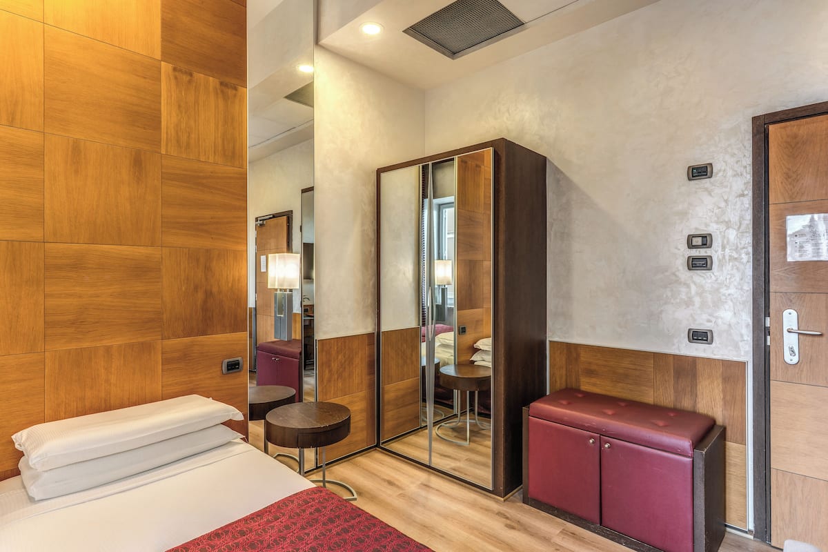 Una habitación de hotel con una cama, una mesita pequeña, un espejo, un armario y un banco rojo.