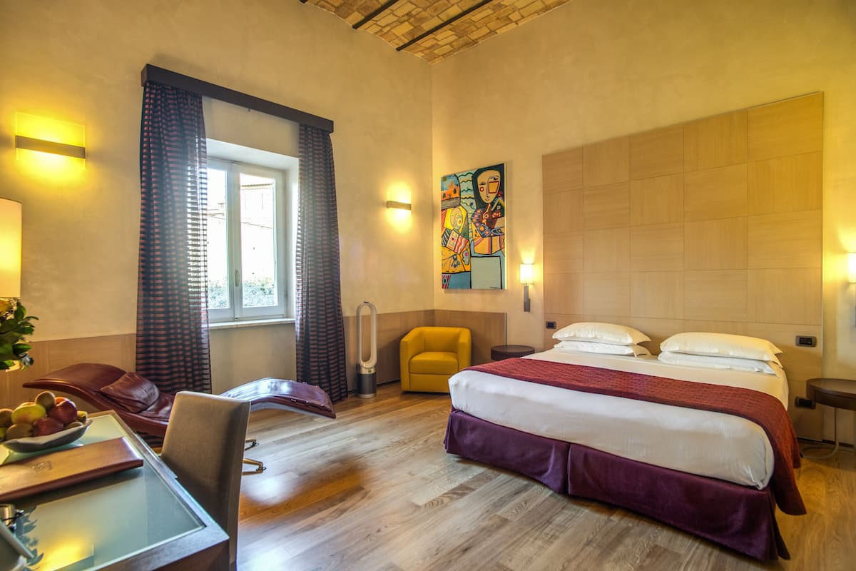Una habitación de hotel con una cama grande, un escritorio con silla, un sillón pequeño, un calentador portátil y un cuadro en la pared.
