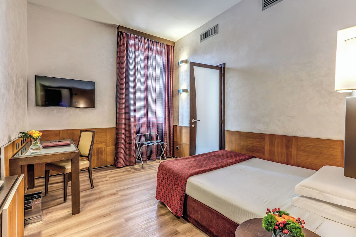 Habitación de hotel con una cama, un escritorio con silla, un televisor montado en la pared y una ventana con cortinas.