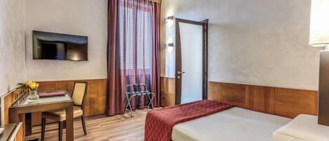 A hotel room with a bed, a desk with a chair, a TV mounted on the wall, and a window with curtains.