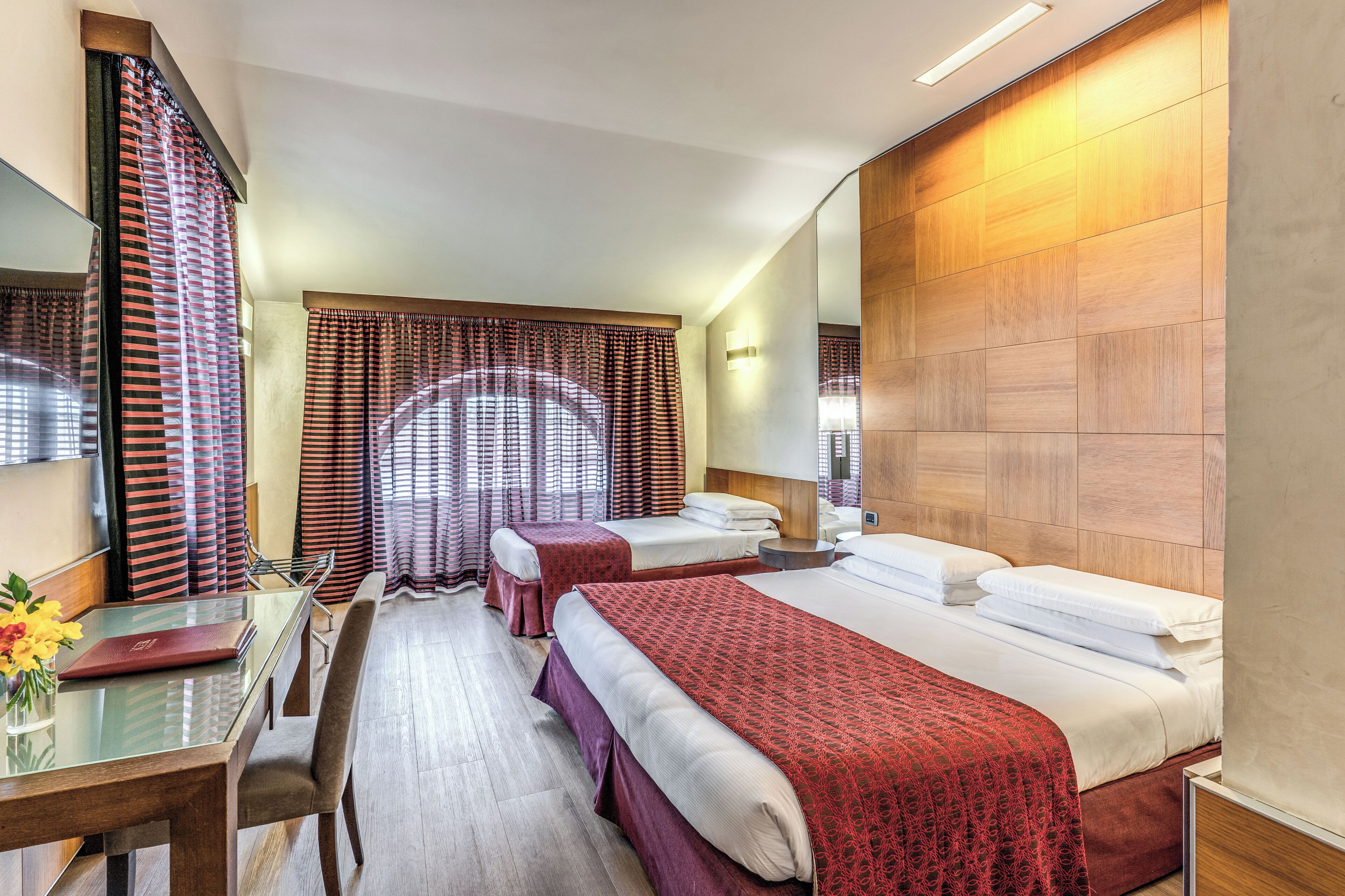 A hotel room with two beds, a dining table with chairs, a mirror, and a window with curtains.