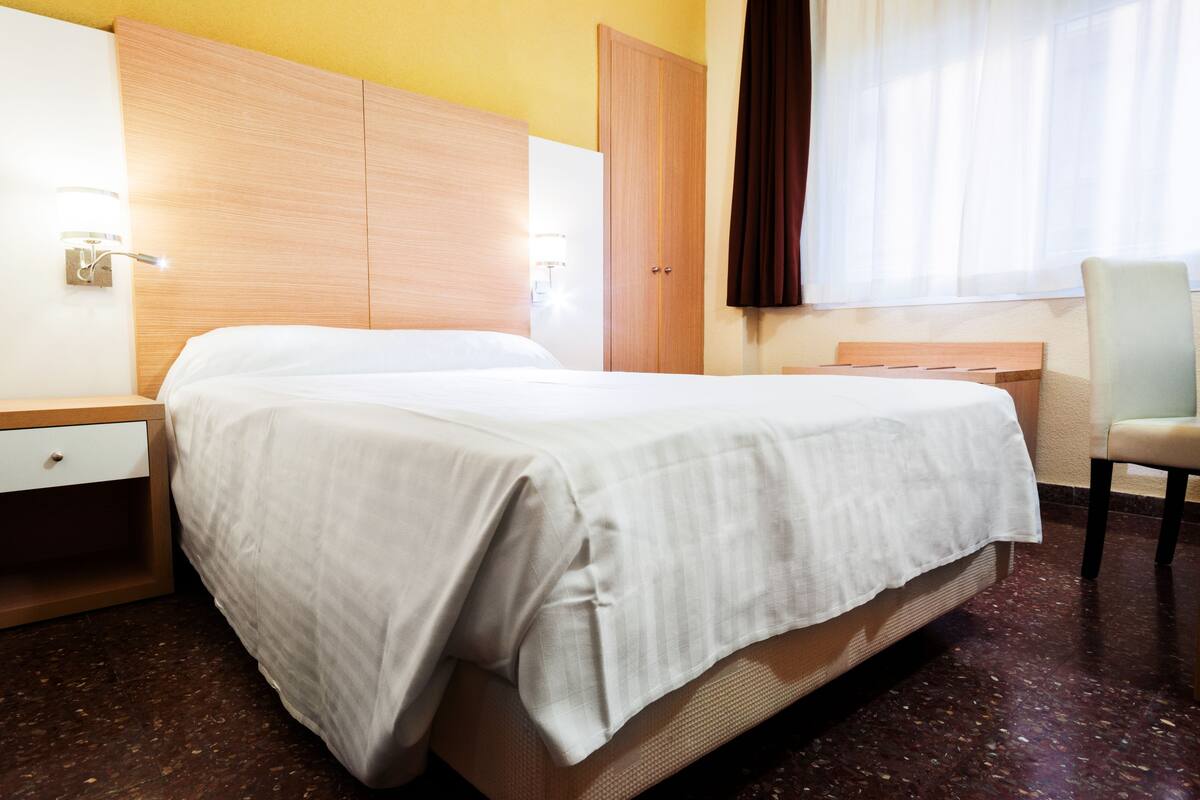 Une chambre d’hôtel avec un lit, une table de chevet, une chaise, une armoire et une fenêtre avec des rideaux.