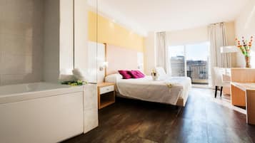 A modern hotel room with a large bed, a separate sitting area, a desk, and a view of the city.