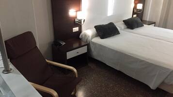 Een hotelkamer met een enkel bed, een stoel, een nachtkastje met een telefoon en een wandlamp.