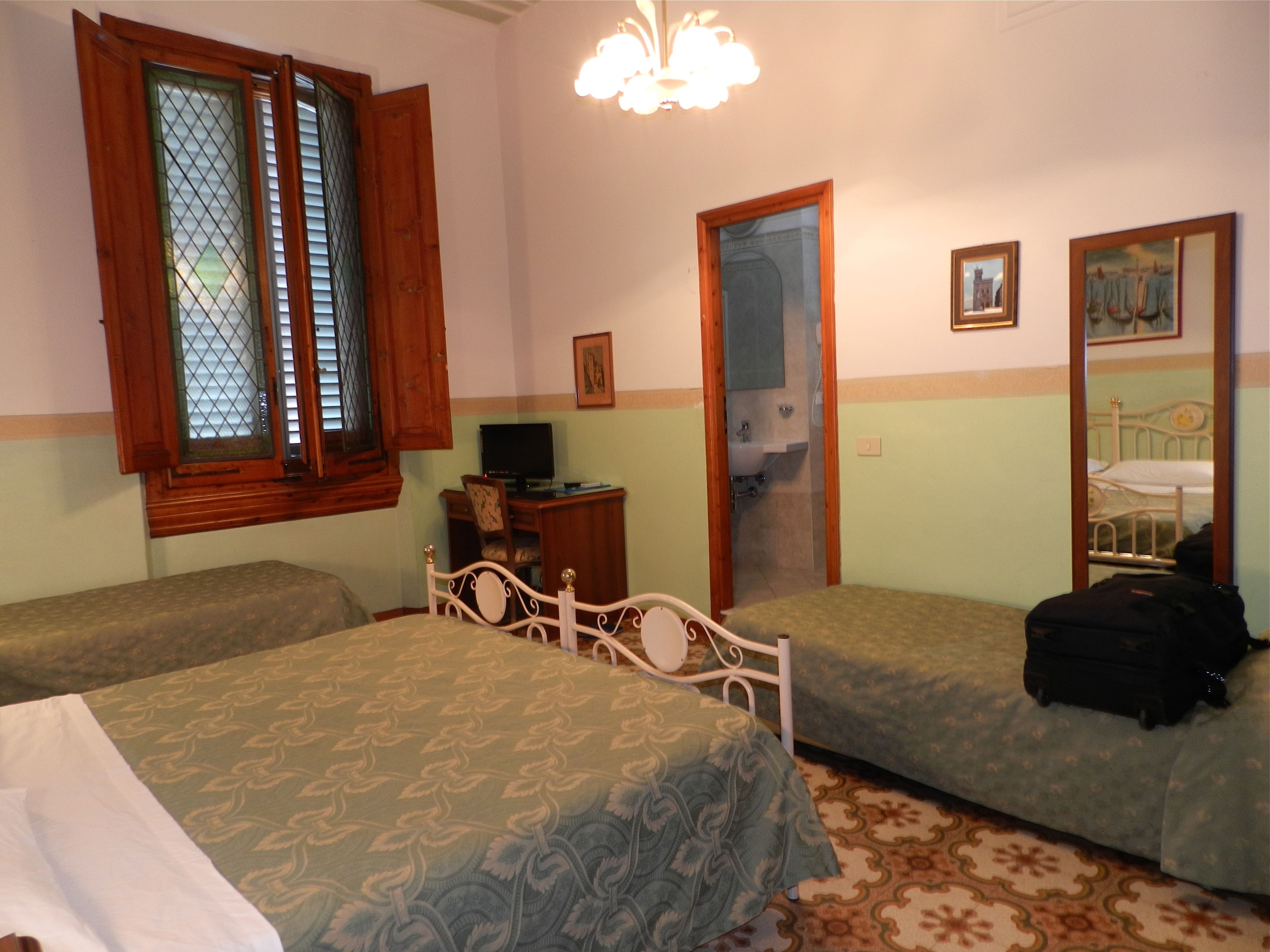 A bedroom with two single beds, a desk with a computer, a mirror, and a window with wooden shutters.