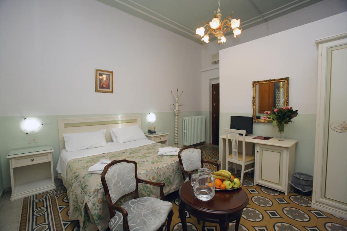 Une chambre d’hôtel comprenant un lit, deux tables de chevet, un bureau avec une chaise, une petite table avec des fruits et un miroir.
