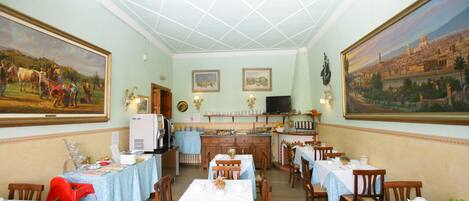 A dining area with tables set for multiple people, a kitchen area with appliances, and two large landscape paintings on the walls.