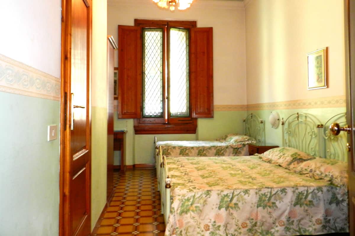 Une chambre avec deux lits, une fenêtre avec des volets en bois, un lustre et un sol carrelé.