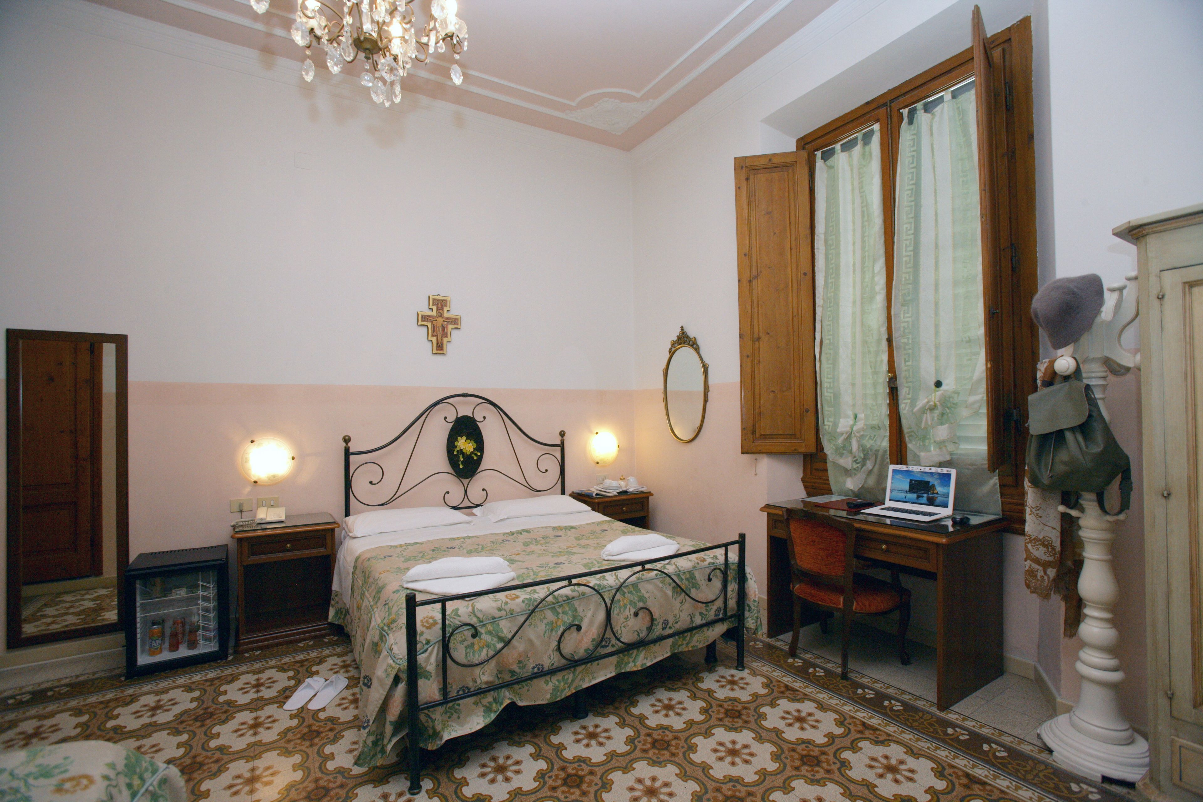 Een slaapkamer met een bed, nachtkastjes, een bureau met een laptop, een stoel, een kledingkast en een kroonluchter.