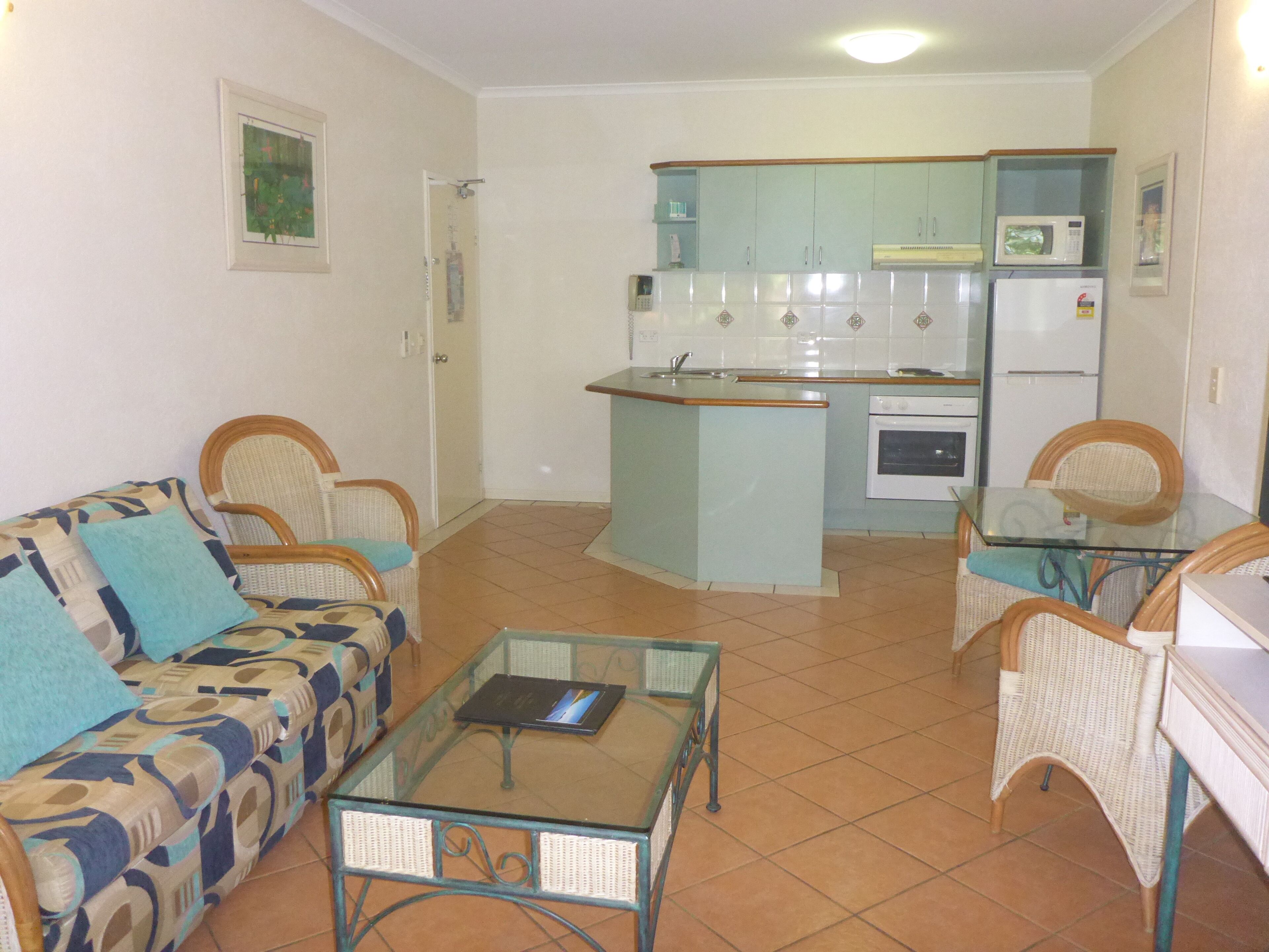A compact living space with a kitchenette, a sofa, a glass coffee table, and wicker chairs.