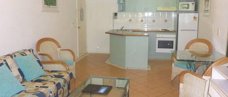 A compact living space with a kitchenette, a sofa, a glass coffee table, and wicker chairs.