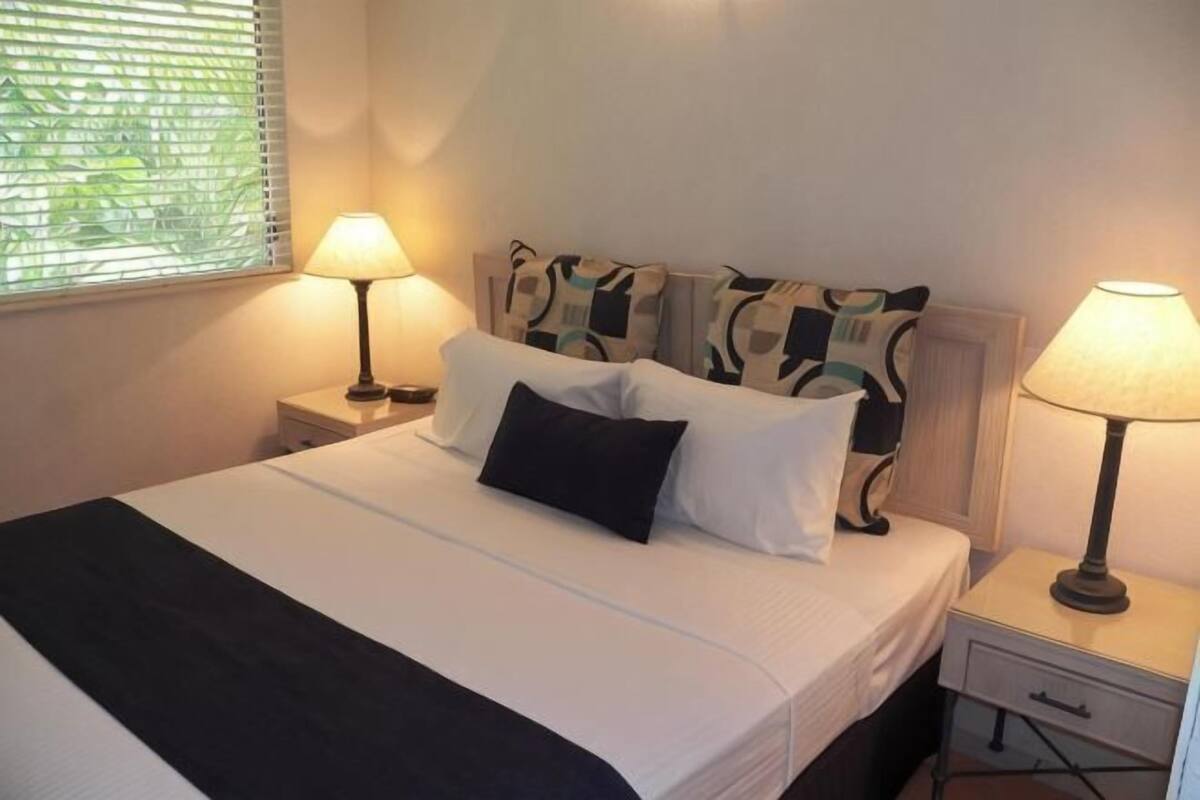 A neatly made bed with pillows, a bedside table with a lamp, and a window with blinds.