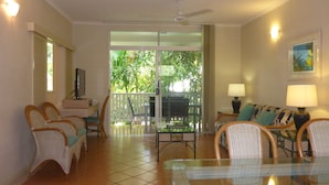 A living room with wicker furniture, a glass table, and a balcony with a view of greenery. - Palm Cove Tropic Apartments (Palm Cove)