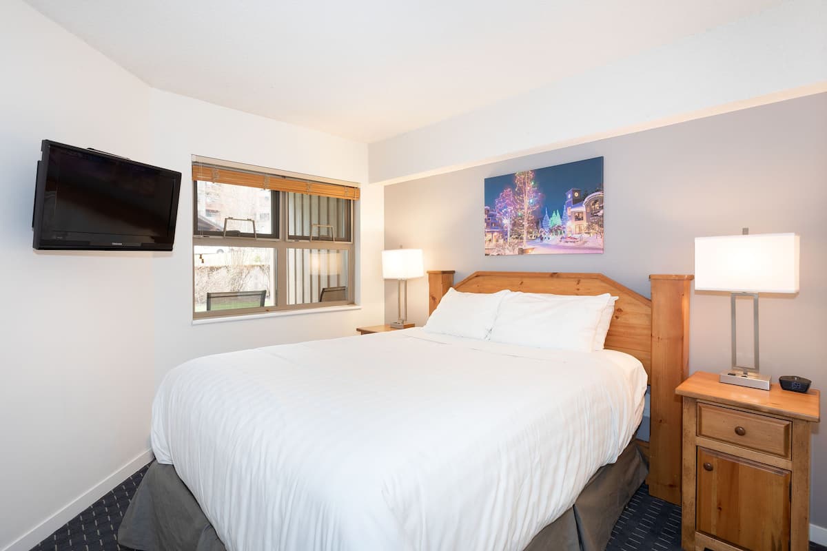 Une chambre d’hôtel avec un grand lit, un téléviseur fixé au mur, une fenêtre avec des stores, une table de chevet avec une lampe et un tableau au mur.