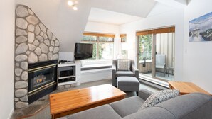 A modern living room with a stone fireplace, grey sofas, a wooden coffee table, and large windows with blinds.