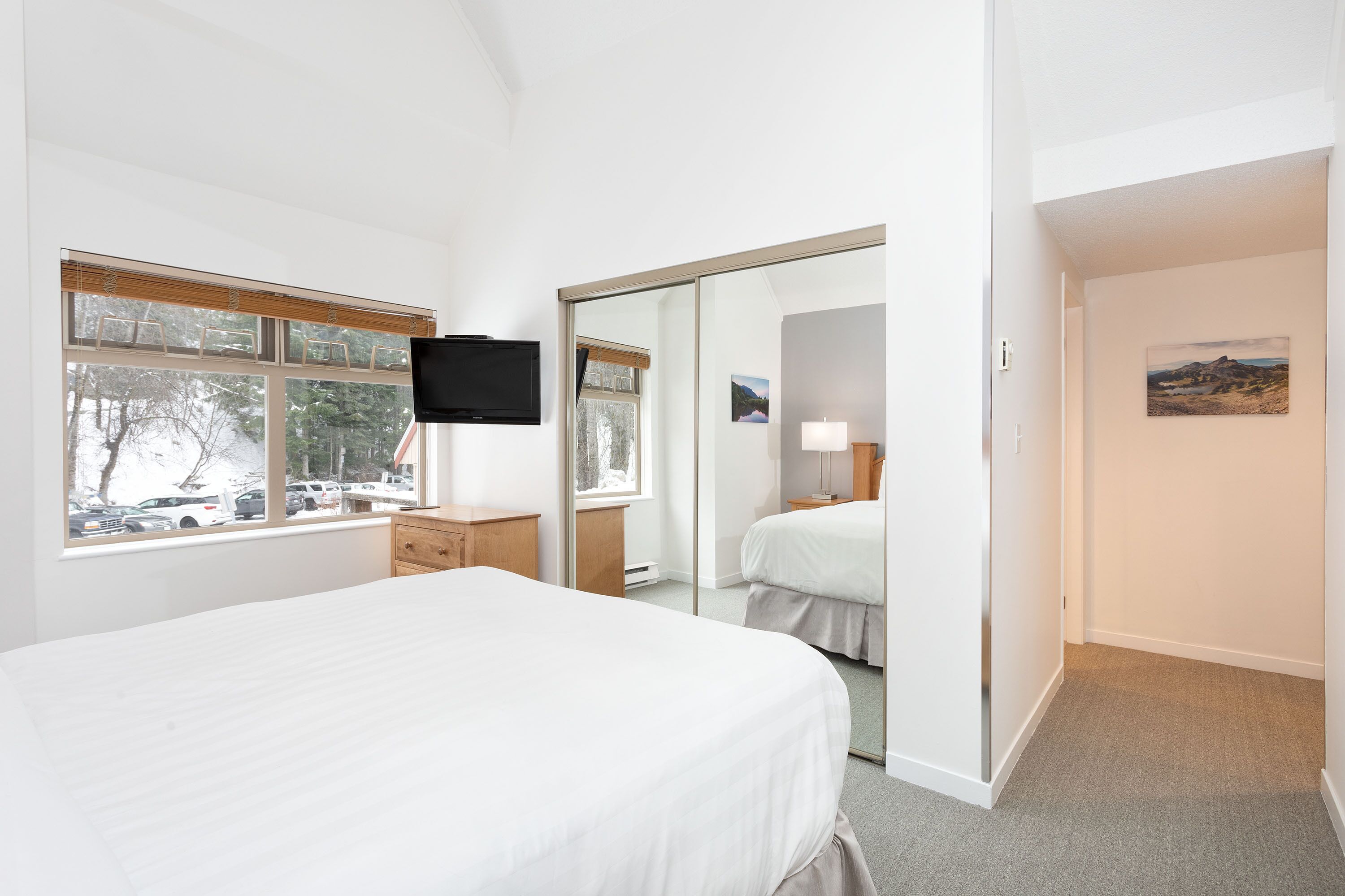 A hotel room with a bed, a TV mounted on the wall, a window with a view of parked cars and trees, and a door leading to another room.