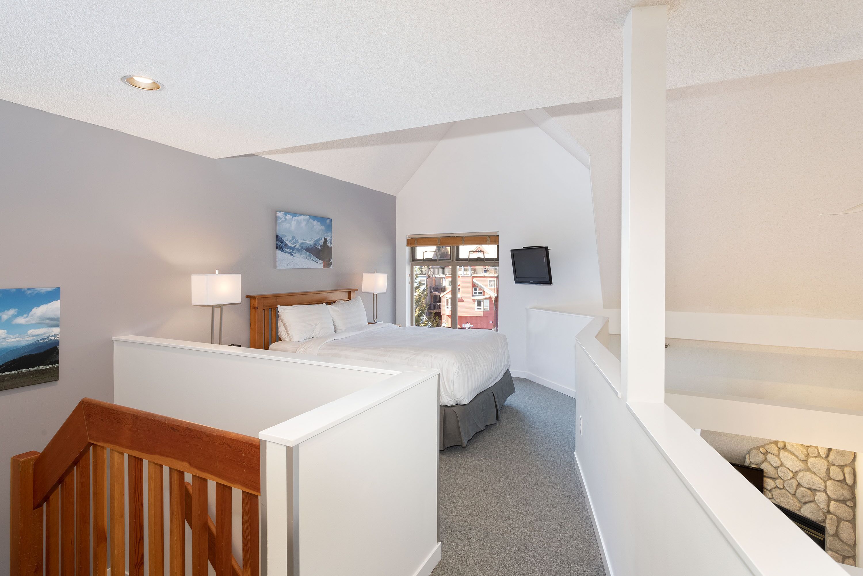 A bedroom with a bed, bedside lamps, a painting, a window, and a TV on the wall.