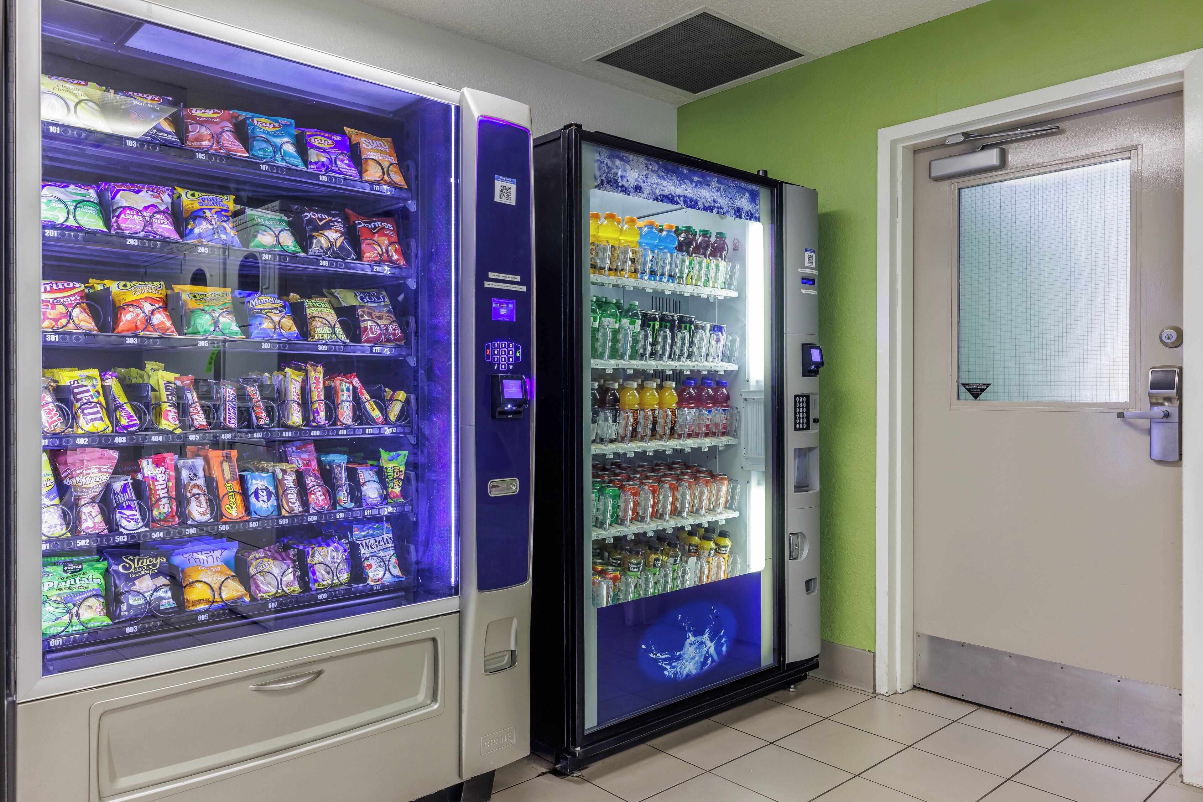 Two vending machines stocked with snacks and beverages in a room with a green wall and a door.