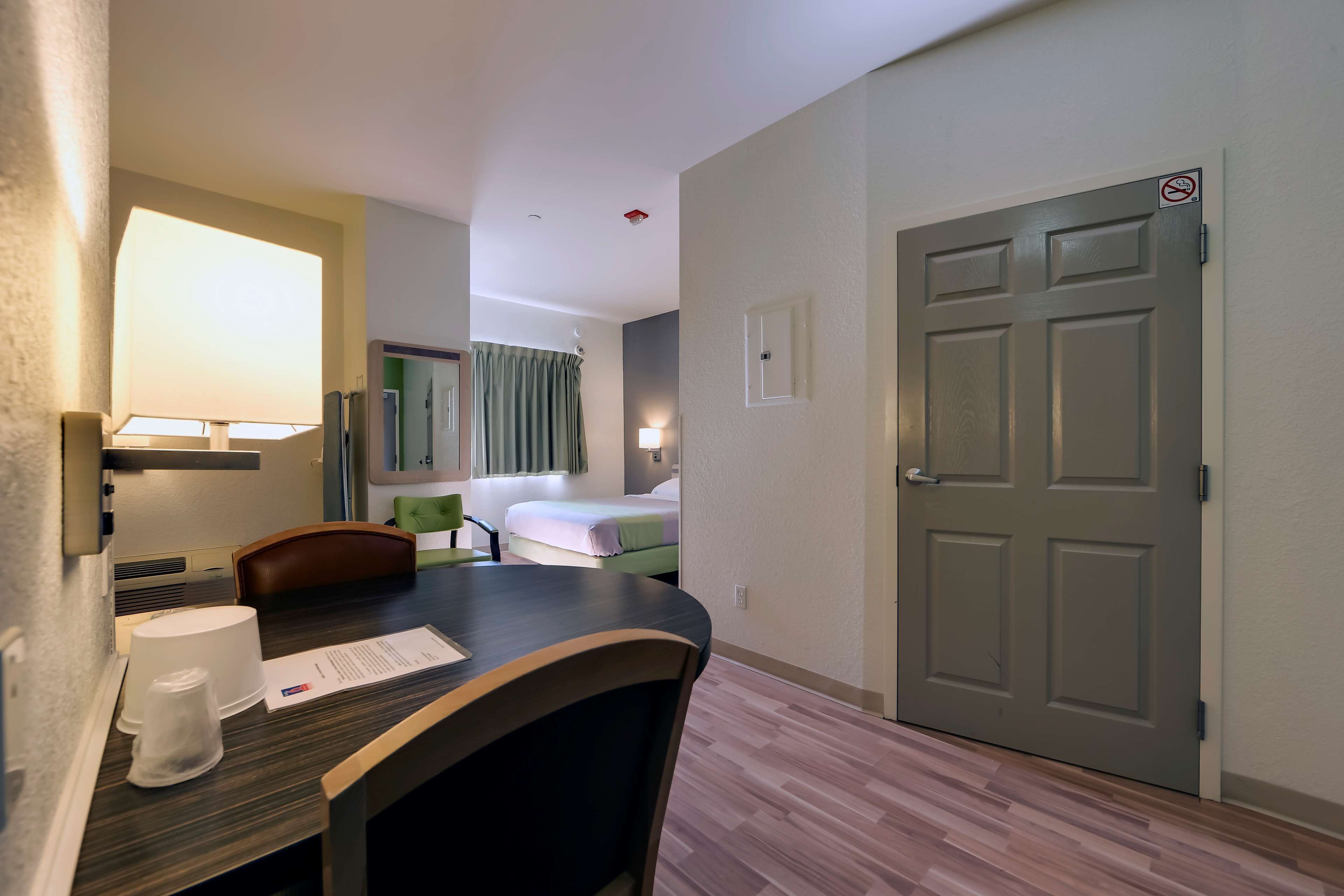 A hotel room with a wooden table, a chair, a bed, a desk, a lamp, a door with a no-smoking sign, and a window with curtains.