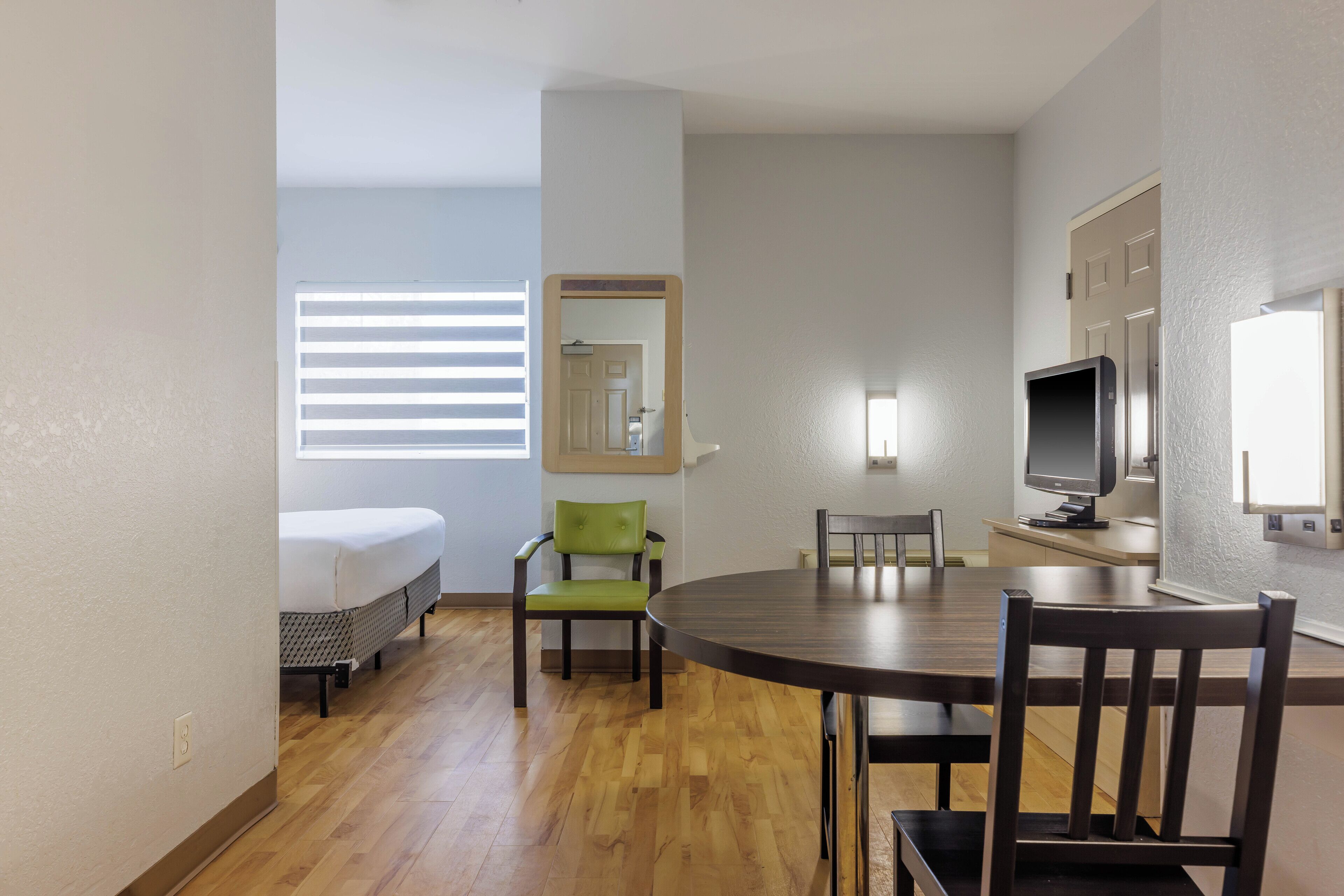 Een hotelkamer met een bed, een groene stoel, een eettafel met stoelen, een televisie en een spiegel.