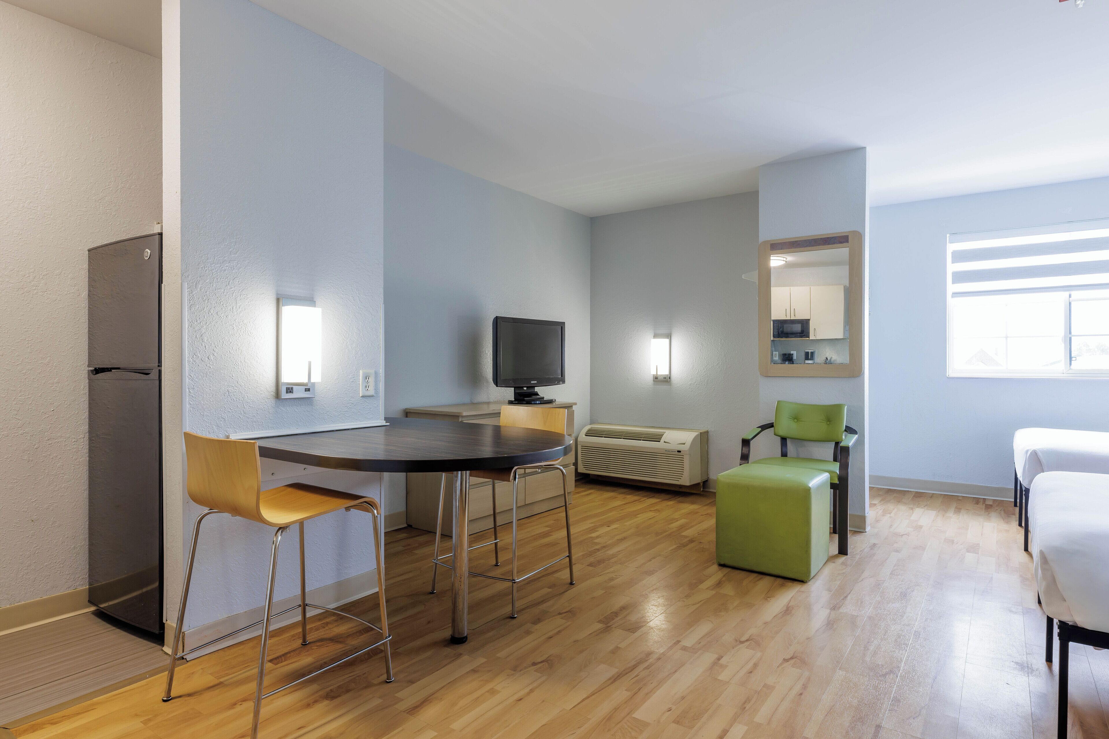 Habitació moderna i ben il·luminada amb un sòl de fusta, una taula de dinar amb caires, una televisió, un mirall i un llit.
