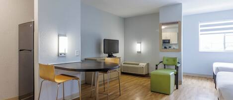 Habitació moderna i ben il·luminada amb un sòl de fusta, una taula de dinar amb caires, una televisió, un mirall i un llit.