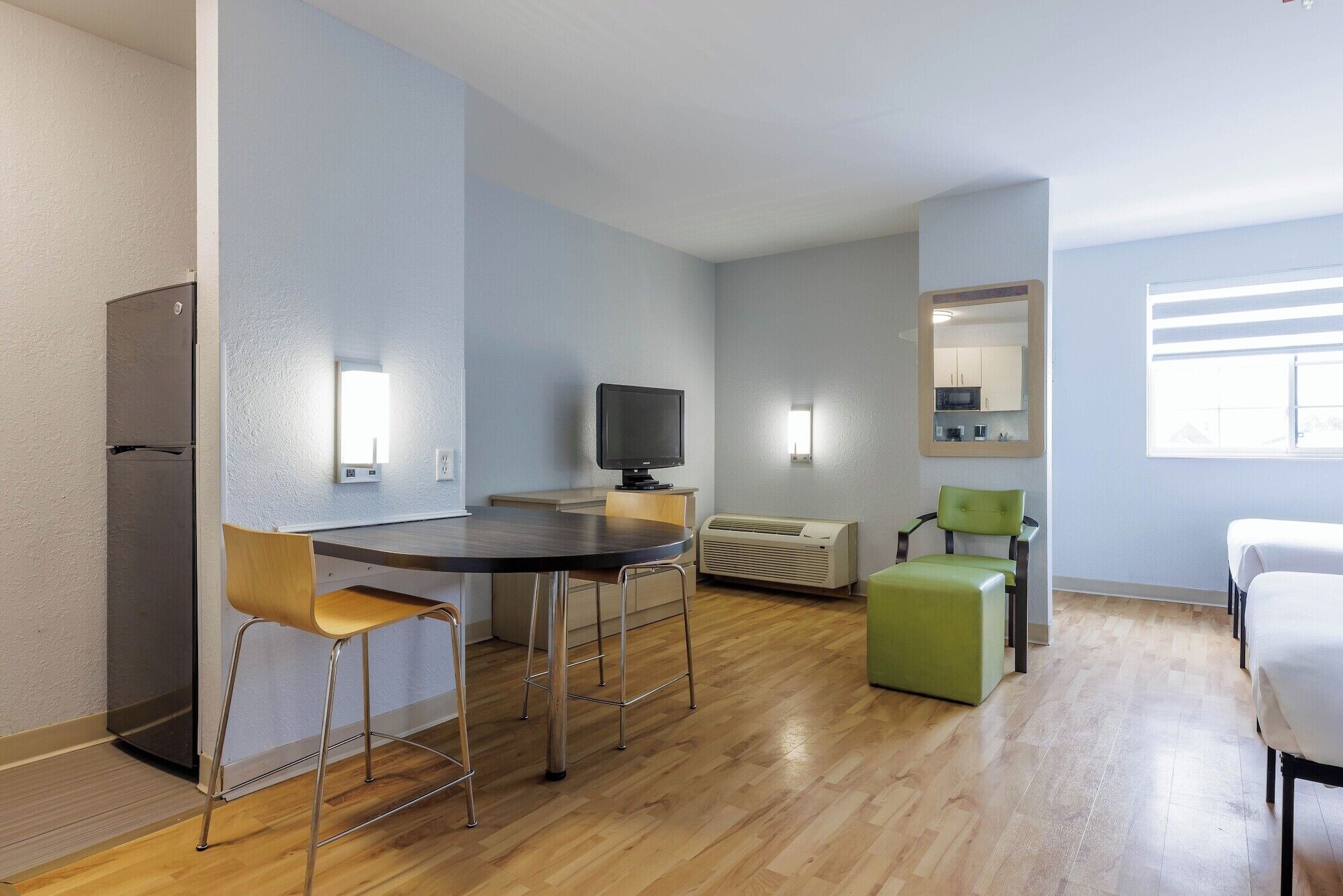 Une pièce moderne et bien éclairée, dotée d’un plancher en bois, d’une table à manger avec des chaises, d’un téléviseur, d’une petite kitchenette et d’un pouf vert.
