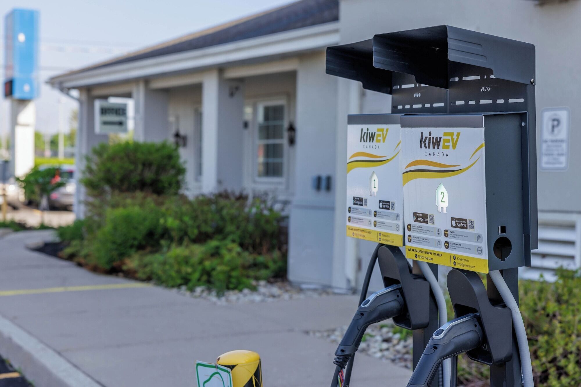 A charging station with two signs for KIWEV Canada, a building with columns, and a parking sign in the background.