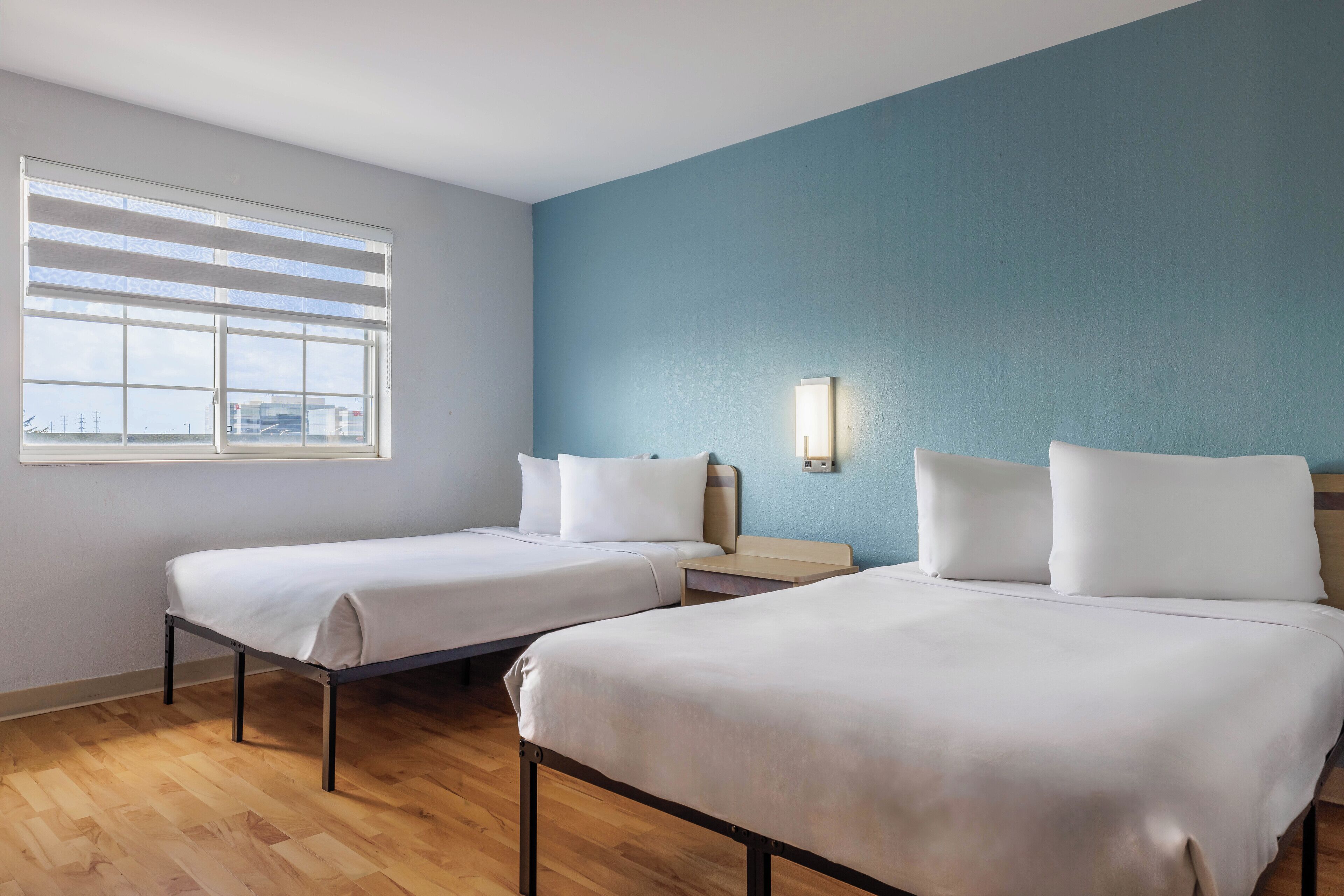 Une chambre d’hôtel avec deux lits, une fenêtre avec des stores, une table de chevet et une lampe fixée au mur.